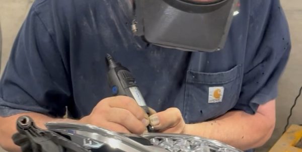Technician using a Dremel rotary tool along the edge of a removed automotive headlamp assembly