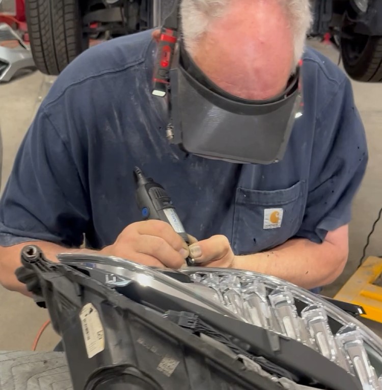 Technician using a Dremel rotary tool along the edge of a removed automotive headlamp assembly