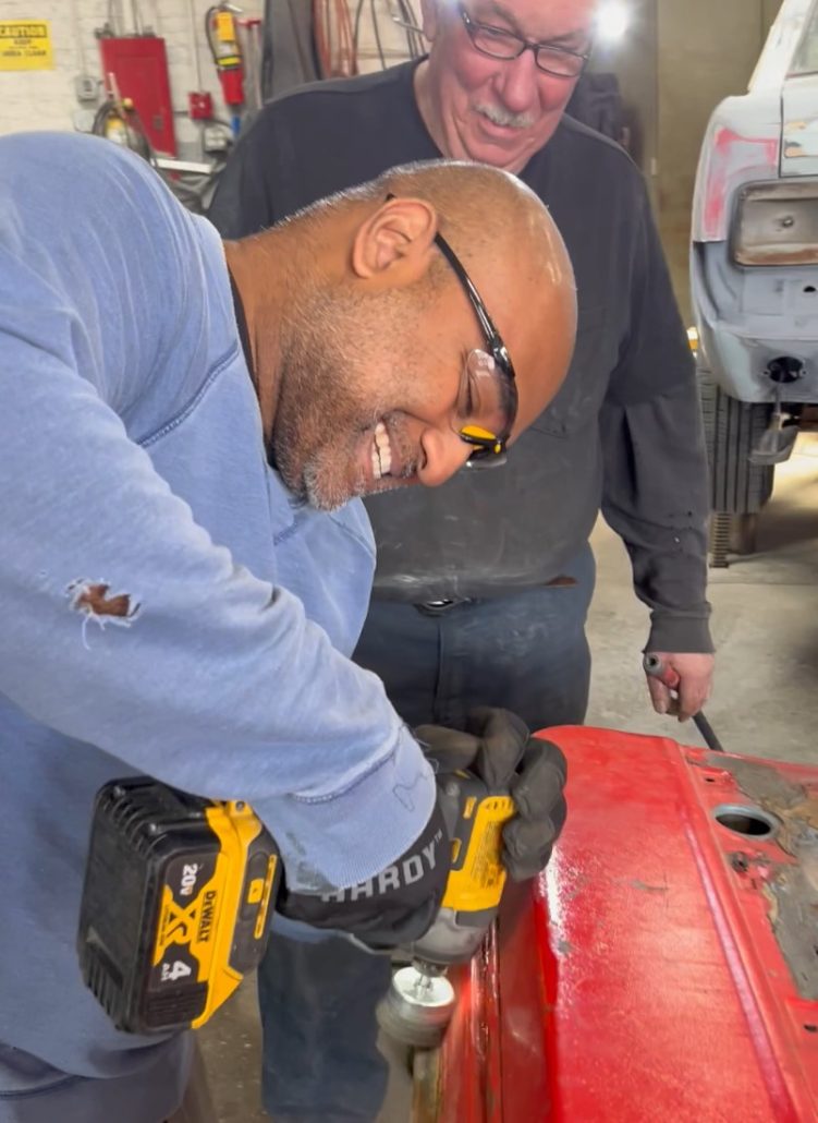 Autoseum student using a drill-mounted wire brush along the edge of a removed red car door