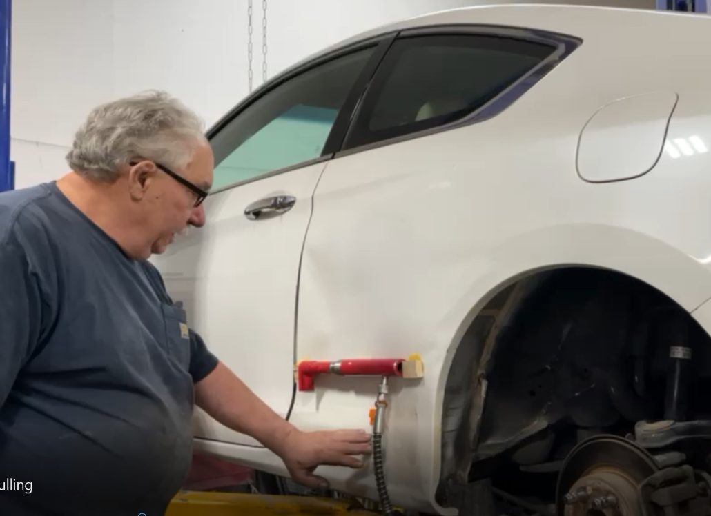Technician working on a white car’s rear quarter panel with red pulling equipment attached to glued wood blocks